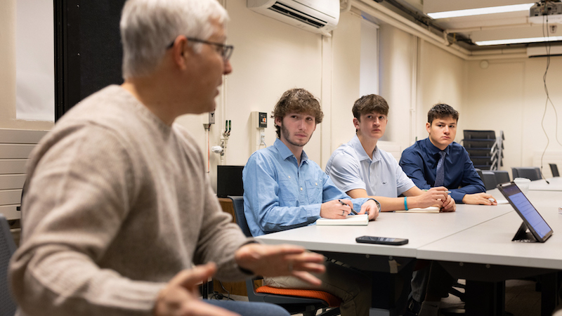 Three students sitting at a table with the professor next to them speaking.