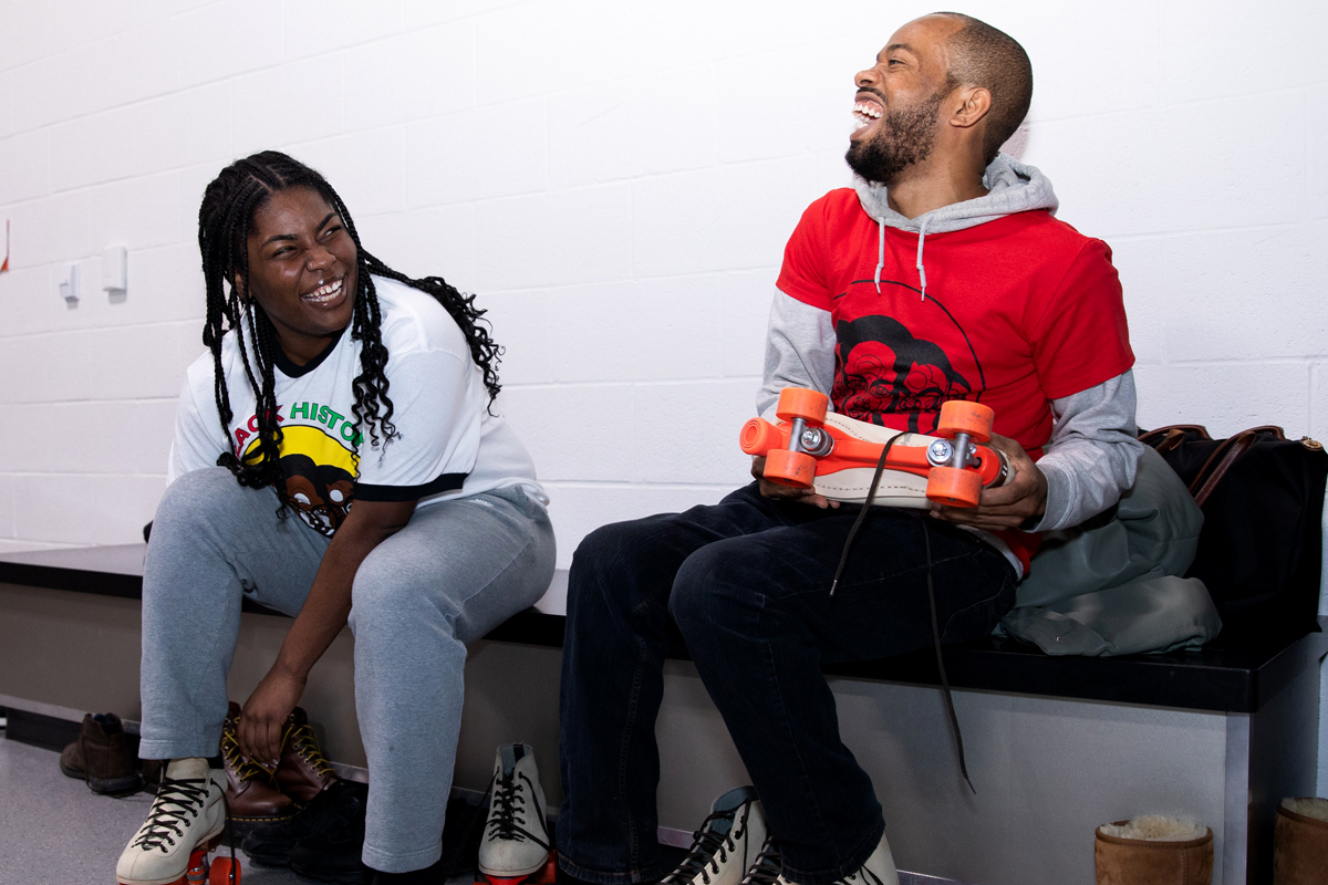 Two people laugh while putting on roller skates.