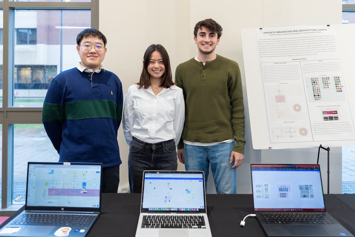 three students stand behind a table with computers displayed on it