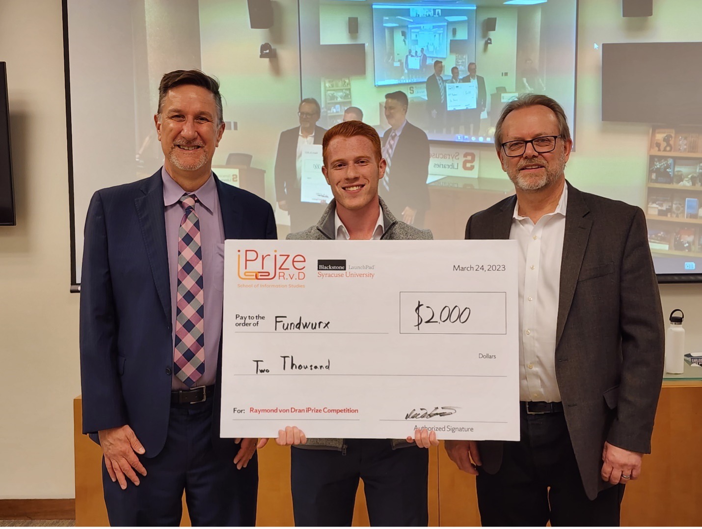 Three people standing together posing for a photo with the person in the middle holding a large check
