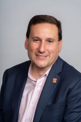 Person in a dark blue suit and pink shirt with an orange “S” lapel pin, against a light gray background.