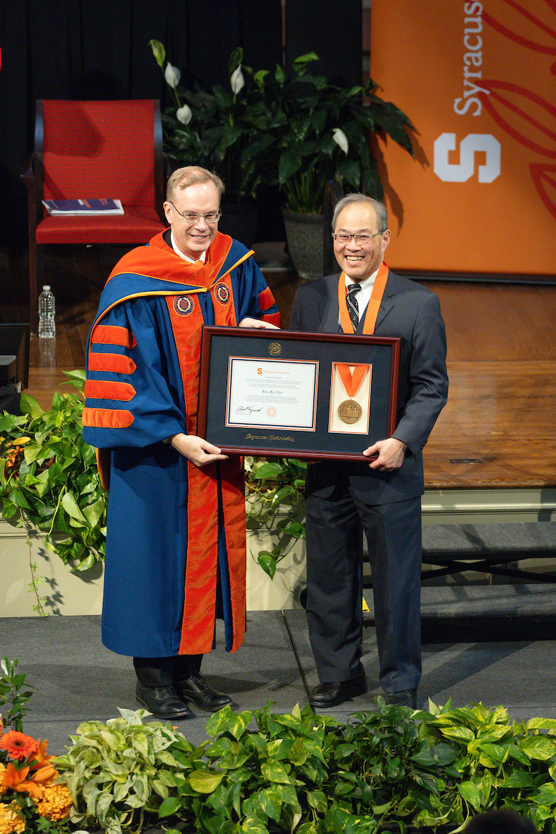 Two people standing together on a stage holding a photo frame with a medal in that. 