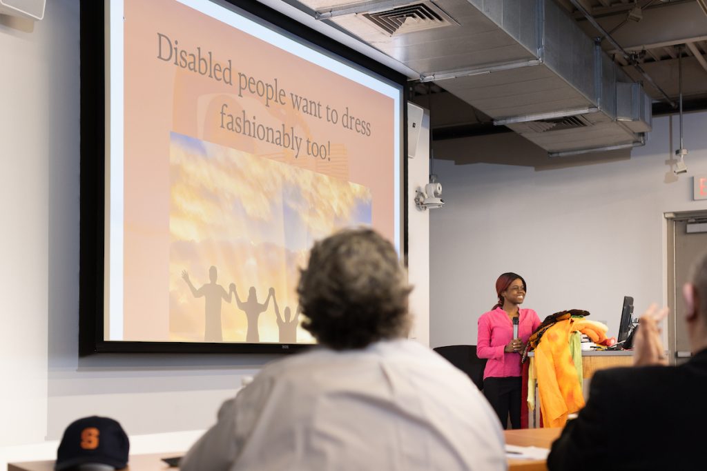 Person standing in the front of a room with a presentation screen behind them speaking into a microphone