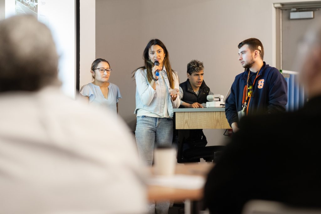 Person in the front of a room speaking into a microphone with three people standing around the individual sepaking