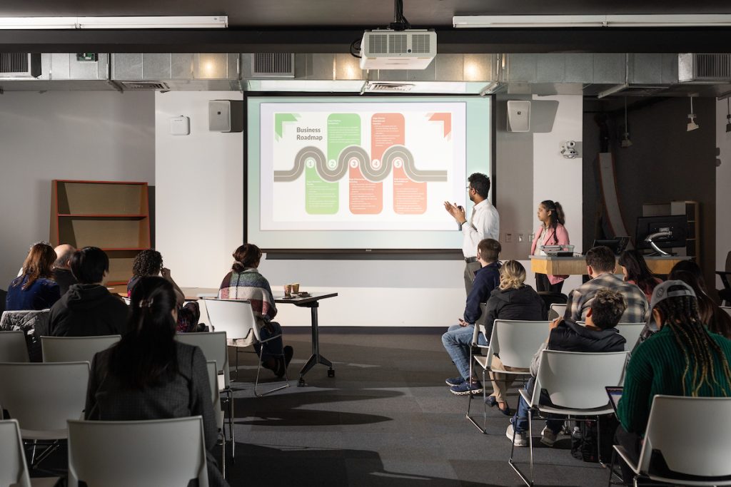 Person standing next to a projection screen speaking to a room of people