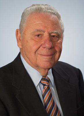 Older person with short white hair in a dark suit and striped tie against a light blue background.