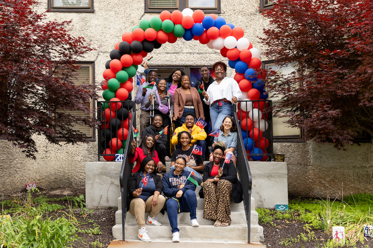 Large group of people gathered on the stairs in front of a stone-sided building with a balloon arch over them with red, black, green, white and blue balloons. 