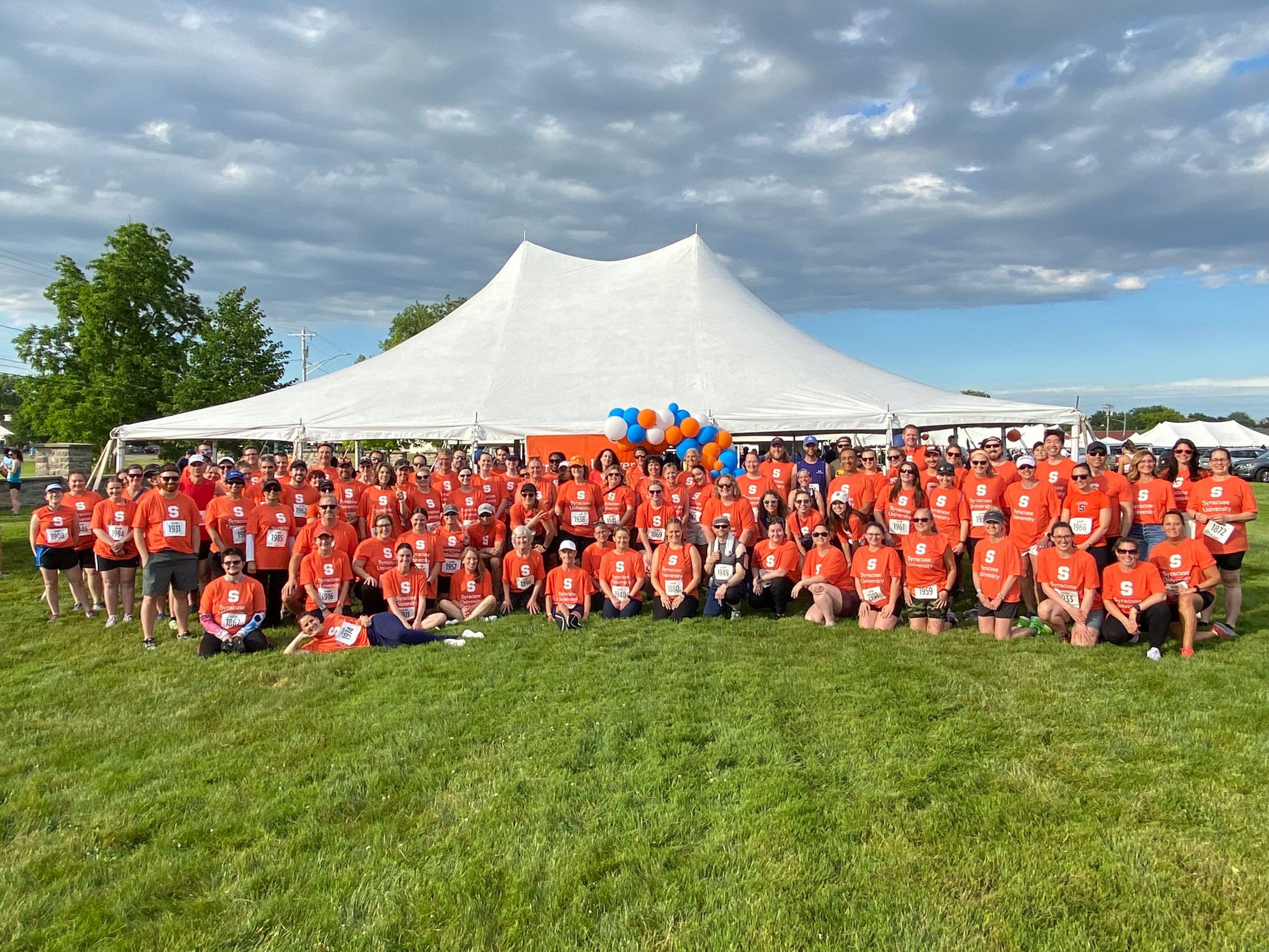 Large group of people gathered together all wearing orange t-shirts outside in front a white tent.