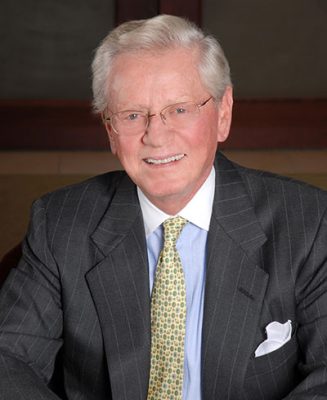 Person in a dark pinstripe suit and patterned tie smiling in front of a blurred background.