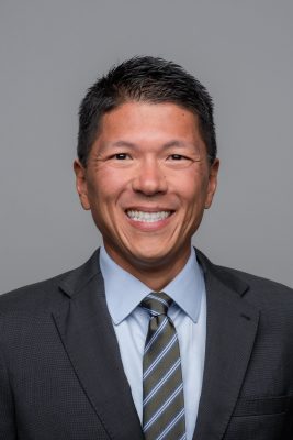 Person in dark suit, light blue shirt, and striped tie smiling against a plain gray background