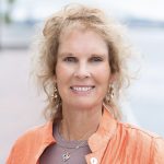headshot of woman with orange coat and purple shirt with water in background