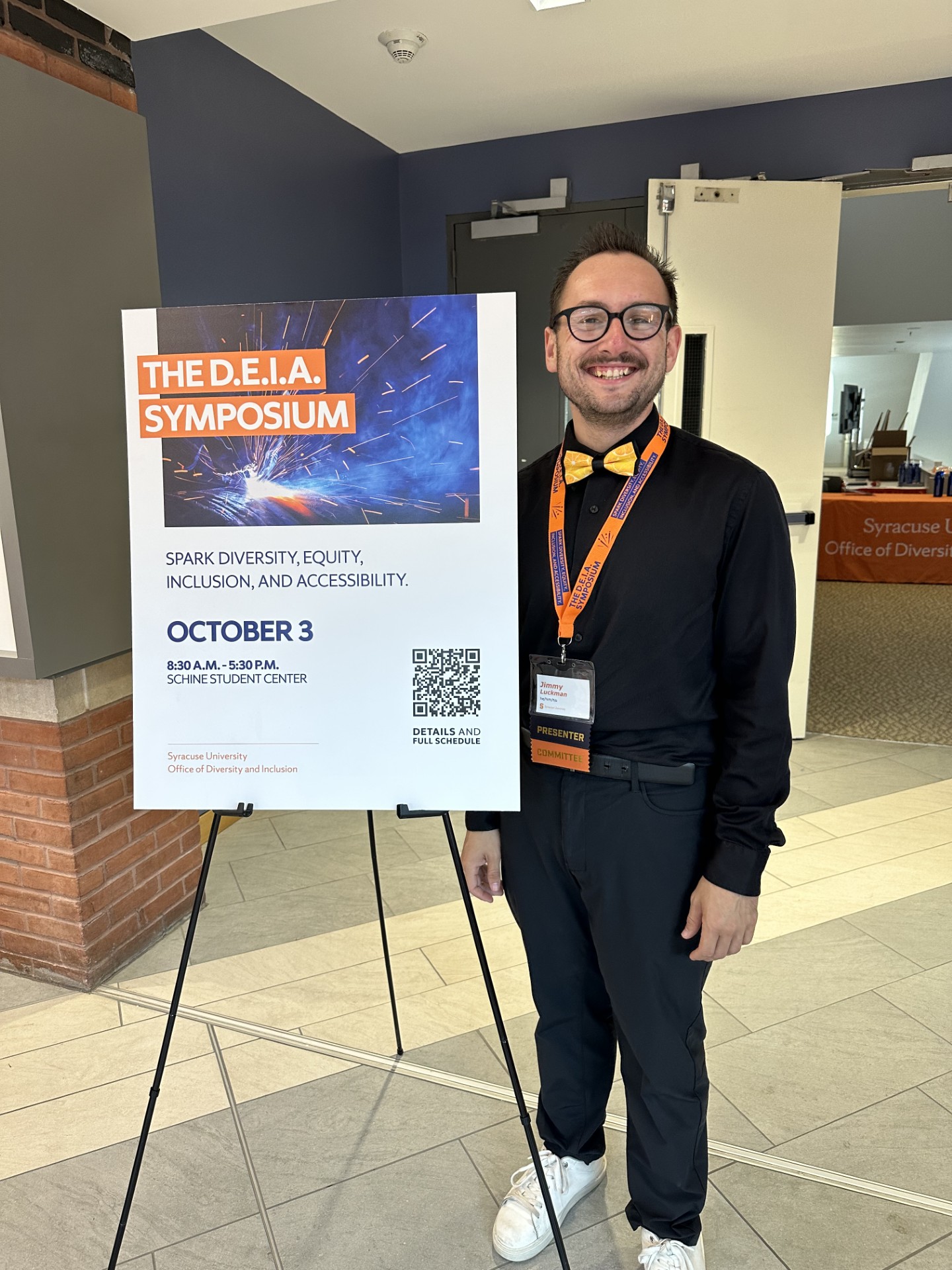 A man smiles for a photo while standing next to a poster for the DEIA Symosium.