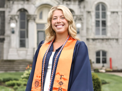 A woman smiles for a photo in front of the Hall of Languages wearing Commencement regalia.