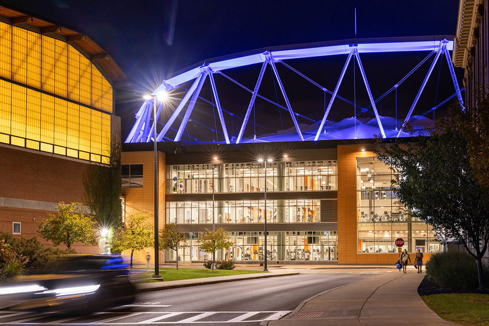 Exterior view of Barnes Center at The Arch lit up and bustling with activity in front of the JMA Wireless Dome roof lit up in blue