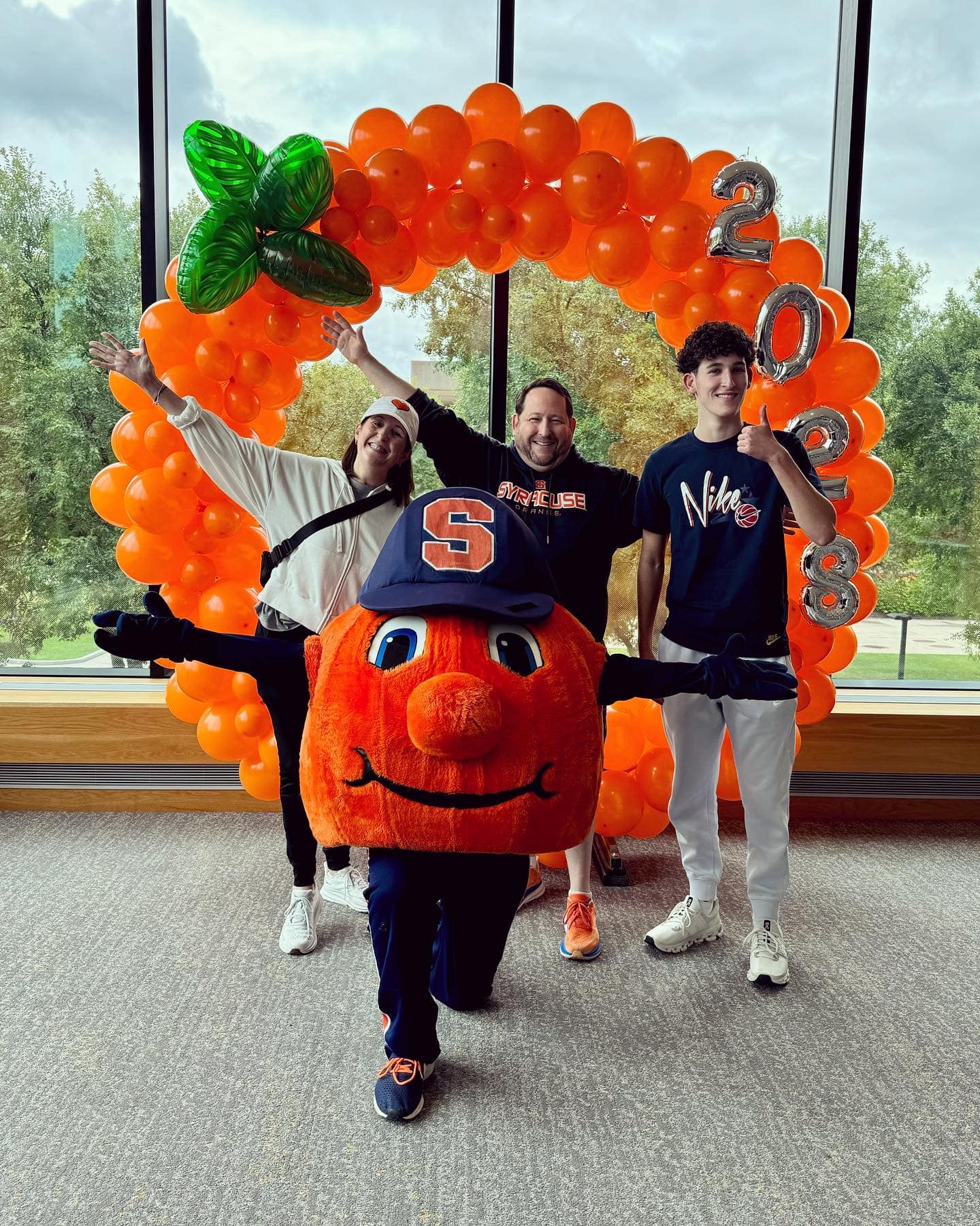 Three people inside with an orange balloon arch and Otto the Orange posing for the photo. 