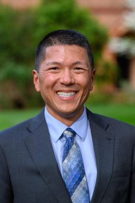 Person in a dark gray suit and blue tie smiling outdoors with greenery in the background.