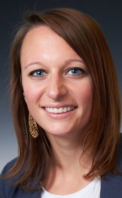 A person smiles while posing for a headshot in front of a grey backdrop.