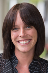 Person with shoulder-length dark hair and bangs, smiling outdoors in a patterned shirt.