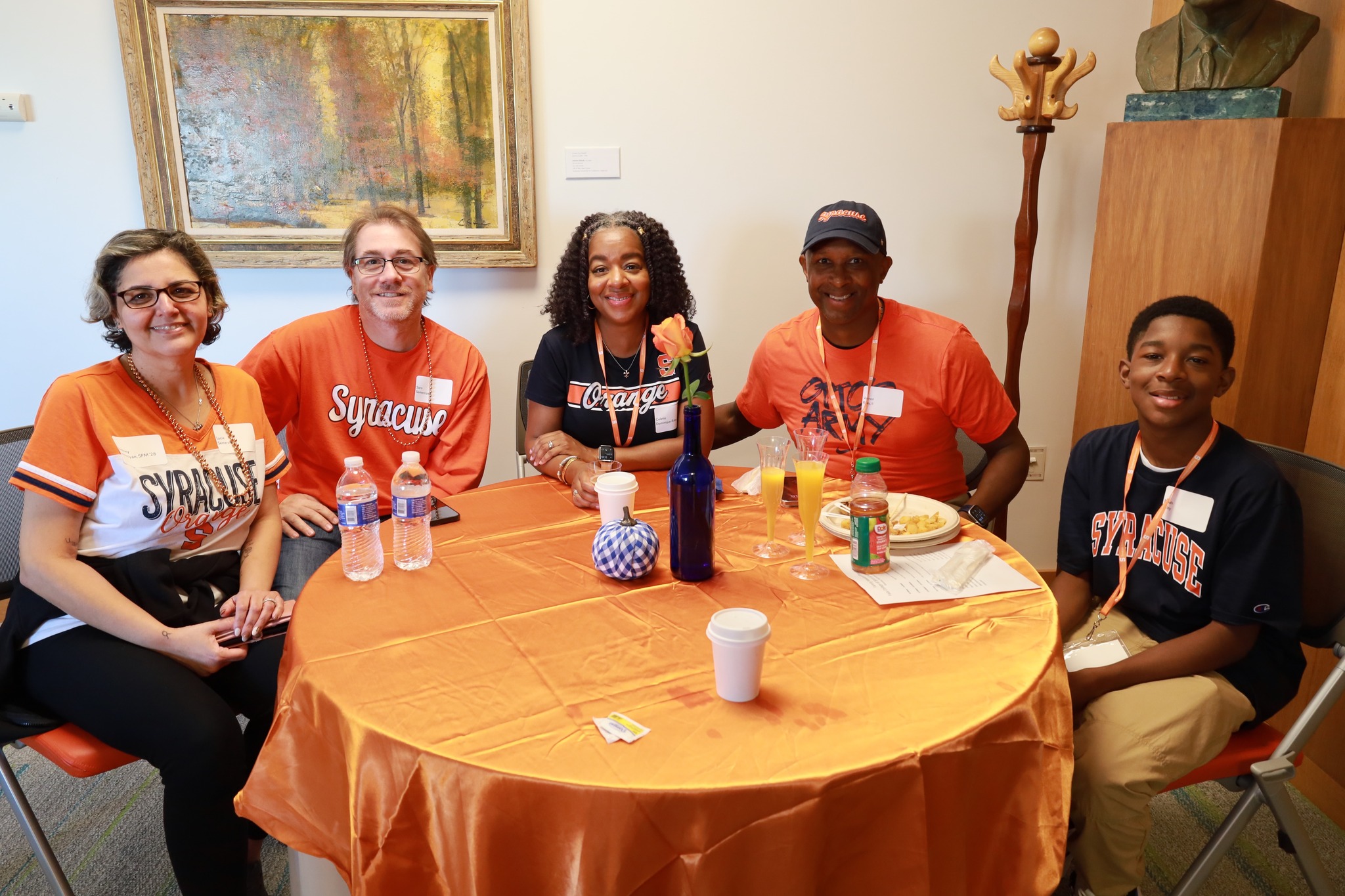 Five people wearing Syracuse University apparel sit around a small table with beverages, smiling at the camera in a room with a painting on the wall.