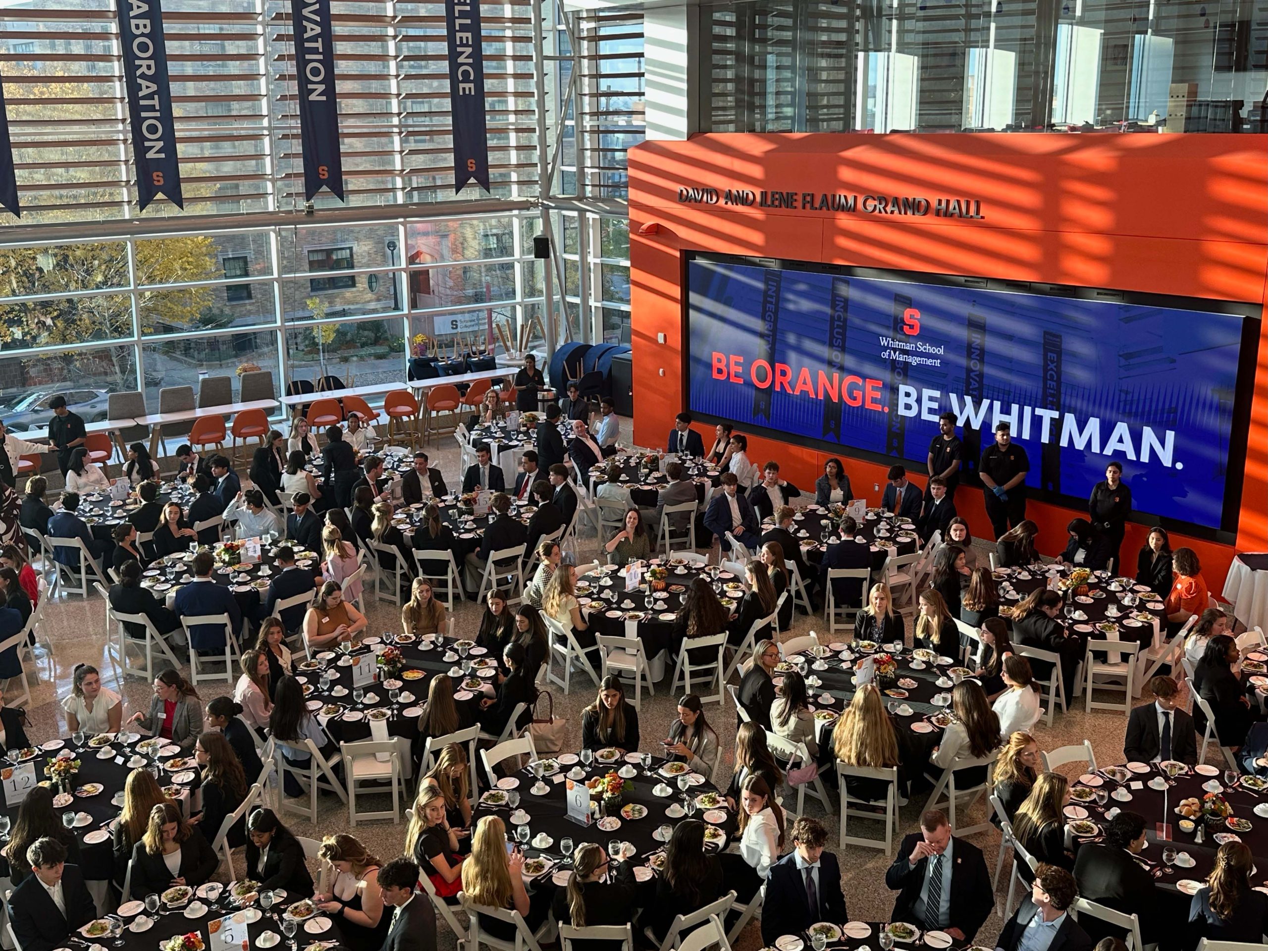Attendees seated at round tables in a large, sunlit hall at the Syracuse University Whitman School of Management event, with a large digital screen displaying school branding in the background.