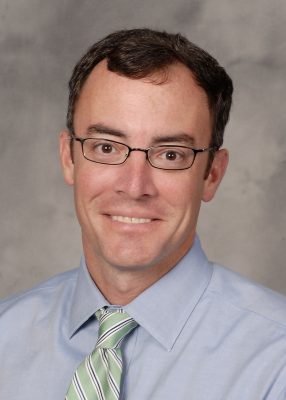 Portrait of an individual wearing glasses, a light blue shirt, and a striped tie, against a grey background.