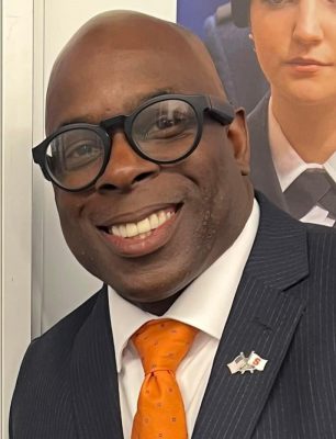 A person smiles while wearing a sport jacket, white dress shirt and orange tie.