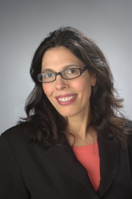 Portrait of a person with long dark hair and glasses, wearing a black blazer over a coral top, against a gray gradient background