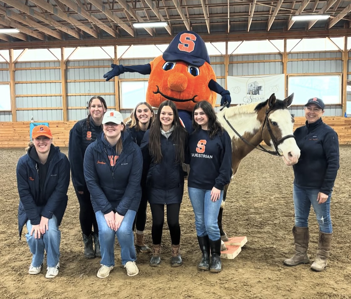 A group of people, some kneeling and some standing, pose for a photo inside an indoor riding arena. They are accompanied by University mascot Otto the Orange wearing a blue cap with a Block S on it. A horse with a white face and brown patches stands next to the group. The individuals are dressed in equestrian attire, including jackets and boots.