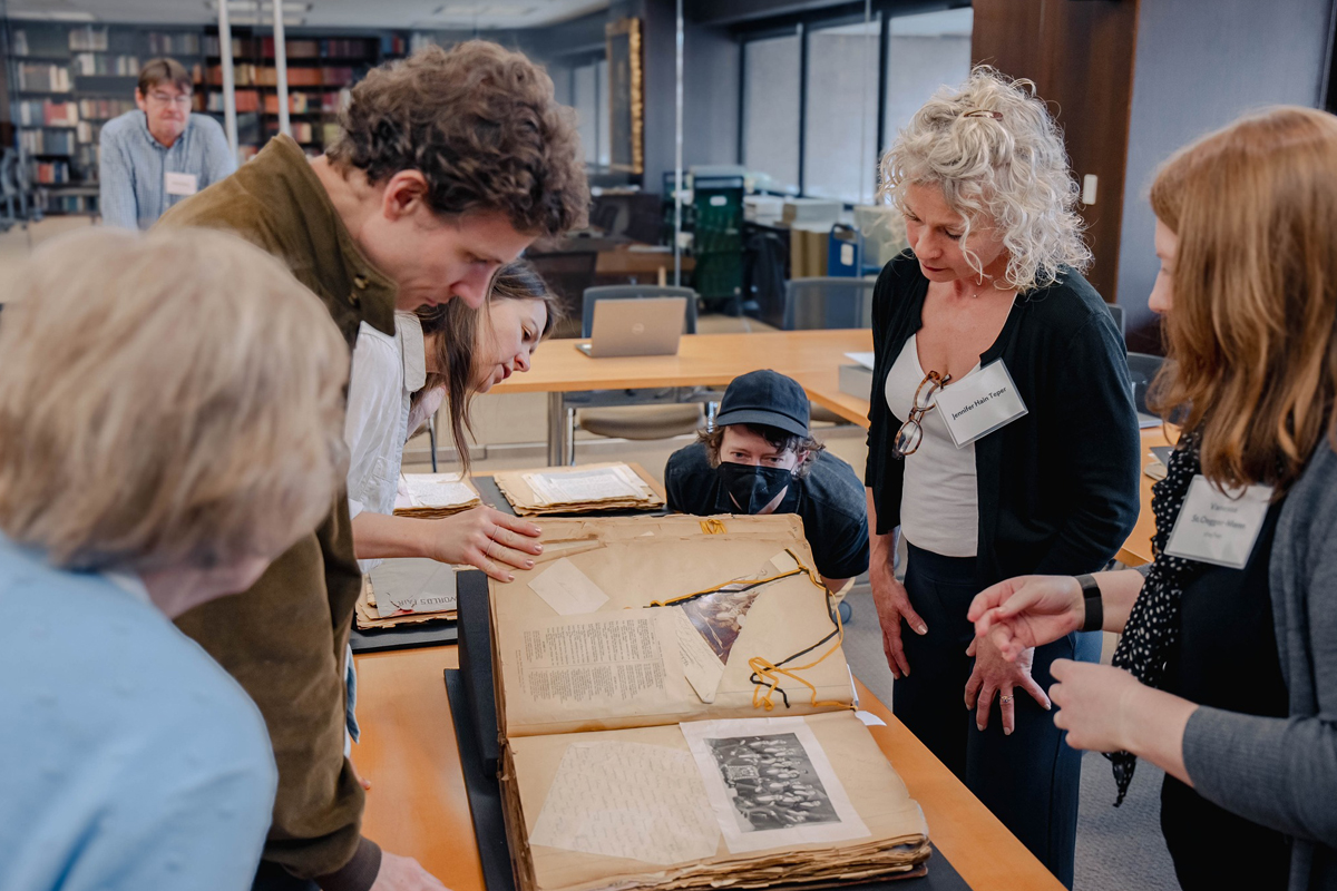 A group of people is gathered around a large, open book on a table in what appears to be a library or archive. The book contains old documents and photographs. The individuals are closely examining the contents of the book, with some leaning in for a better view. Shelves filled with books and other materials are visible in the background.
