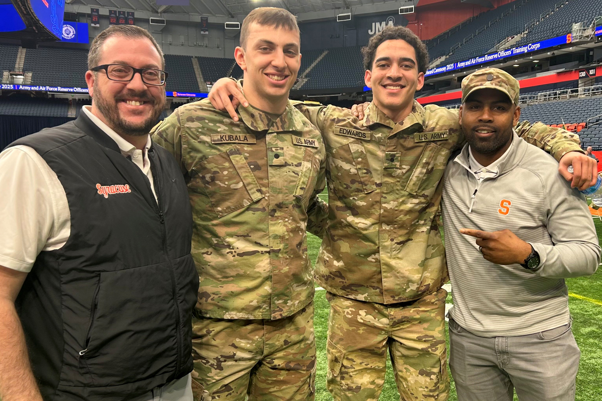 The image shows four individuals standing together in a stadium. Two of them are wearing military camouflage uniforms, while the other two are dressed in civilian clothing. The person on the far left is wearing a white shirt with a black vest, and the person on the far right is wearing a gray shirt and cap, pointing towards one of the individuals in uniform. The background shows empty stadium seats and some banners.