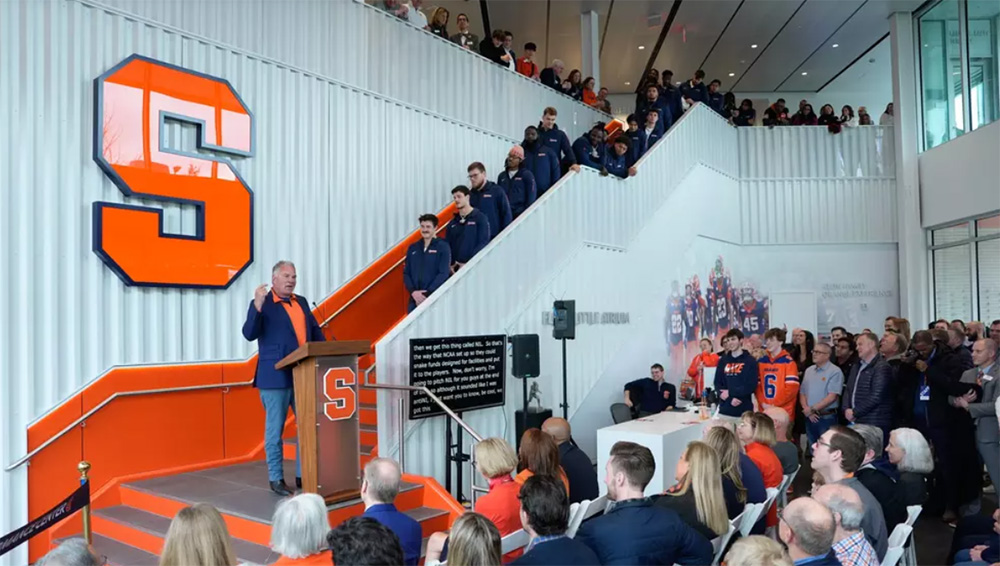 John Lally speaks at a podium with a large Block S on the wall behind him. Several people are standing on stairs in the background, and an audience is seated in front of the speaker. There is a prompter with text next to the podium.