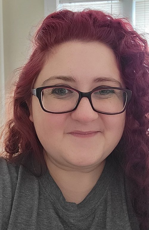 The image shows a person with curly, dark red hair wearing glasses and a gray shirt. The background features a window with blinds partially open, allowing light to enter the room.