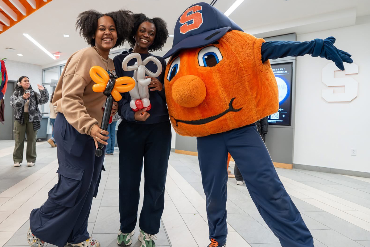 Two people are posing with a large orange mascot wearing a blue cap and blue pants. The mascot has an orange face with a big smile and is holding out its arms. The two people are holding balloon sculptures, one shaped like a flower and the other like a butterfly. They are standing in what appears to be an indoor hallway or lobby area with tiled floors and white walls. There is another person in the background on the left side of the image, partially visible.