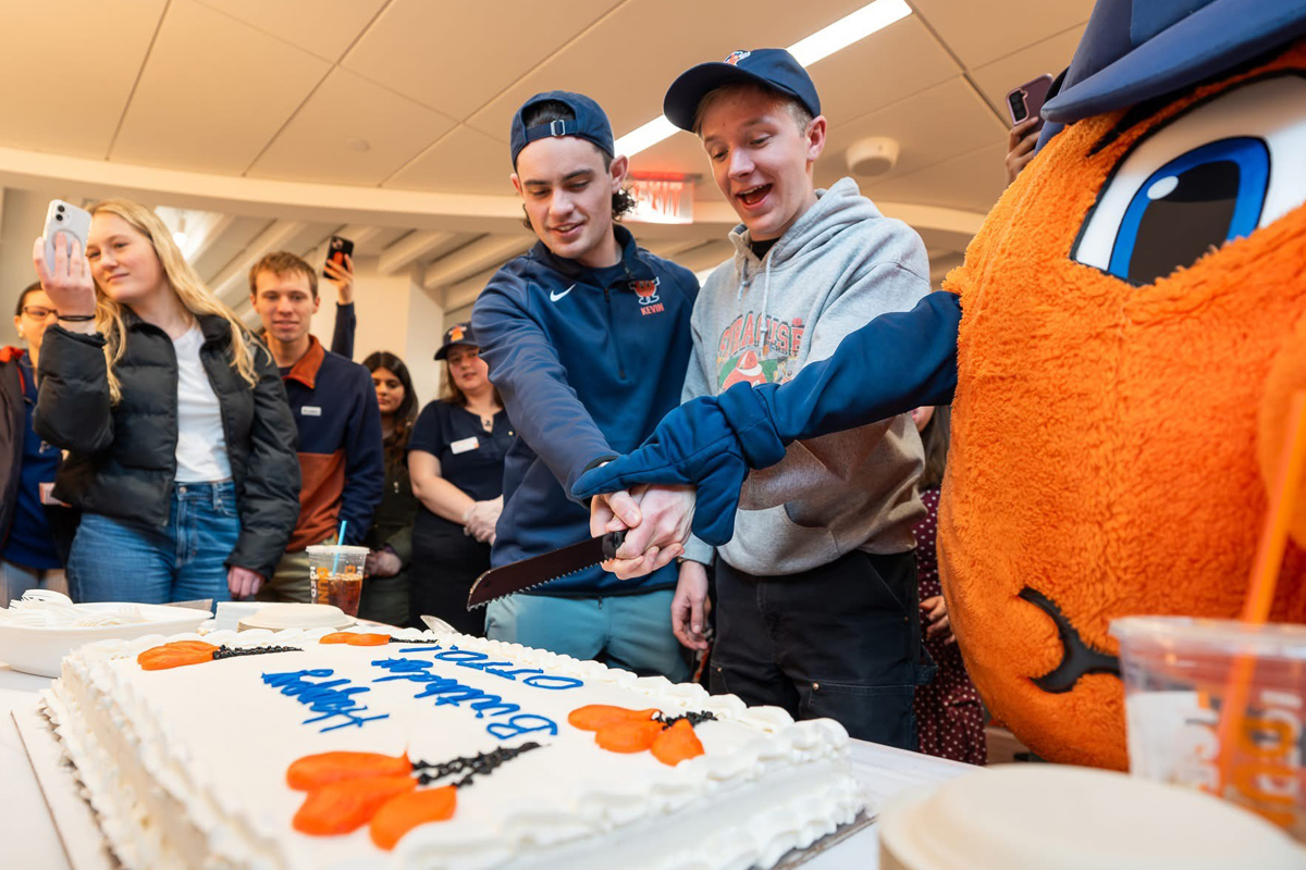 A group of people is gathered around a large rectangular cake with white frosting and blue and orange decorations. Two individuals in the center are cutting the cake together, holding a knife. To the right, there is a person in an orange mascot costume with large eyes. Several people in the background are taking photos or observing the scene. The setting appears to be indoors, possibly at an event or celebration.