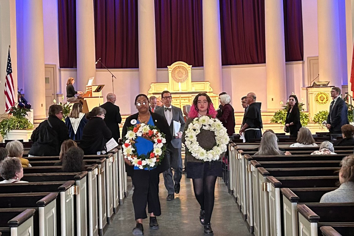 The image shows an indoor ceremony with people walking down the aisle of a church or similar venue. Two individuals in the foreground are carrying large wreaths made of white flowers, with one wreath also containing red and blue flowers. The background features tall columns, a stage with an organ, and several other people standing or walking. The setting appears formal, with attendees dressed in suits and dresses.