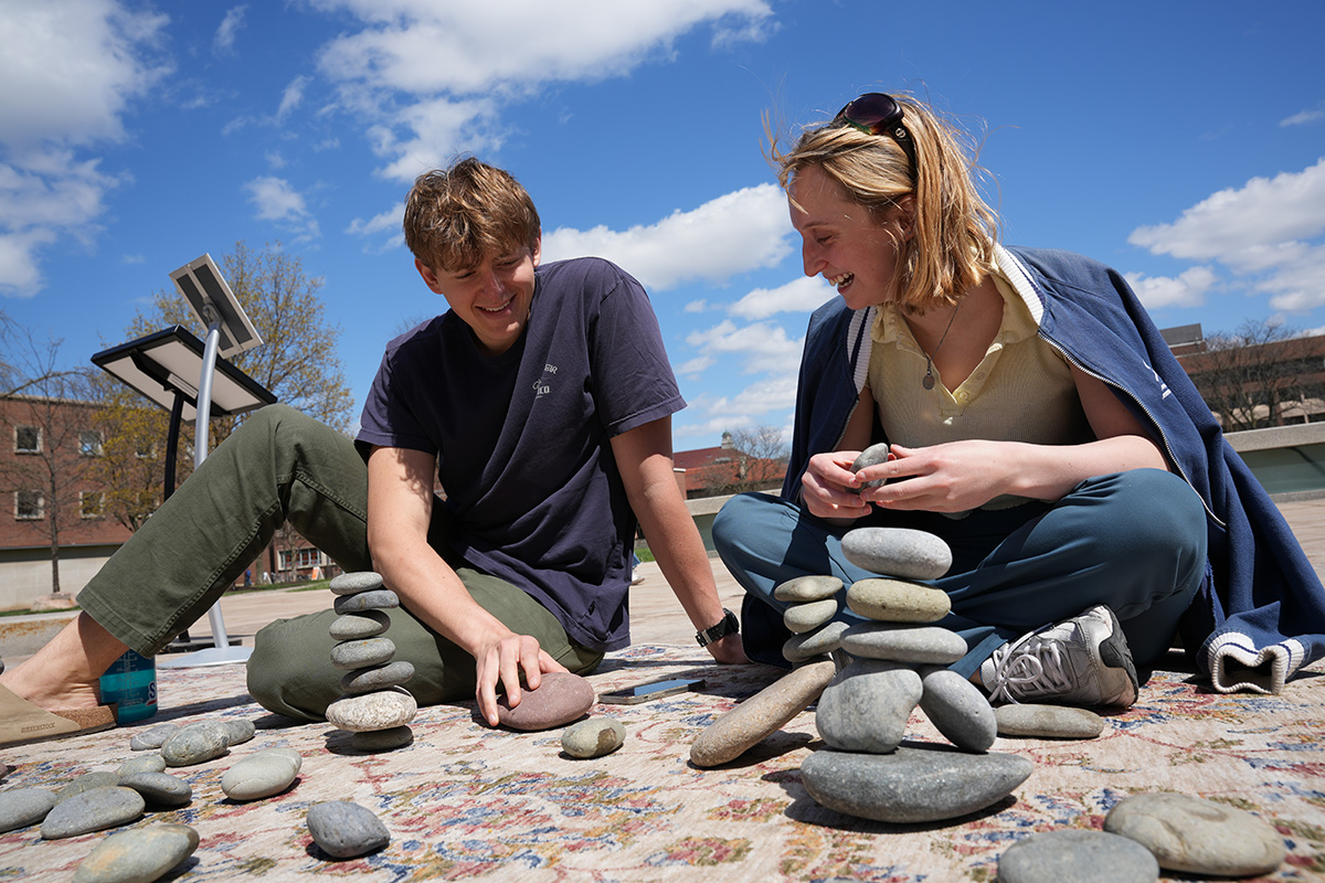 Two people are sitting on a patterned rug outdoors, under a blue sky with scattered clouds. They are engaged in stacking smooth, flat stones into small towers. The person on the left is wearing a dark shirt and green pants, while the person on the right is wearing a light-colored shirt and blue pants with a jacket draped over their shoulders. In the background, there are buildings and trees.