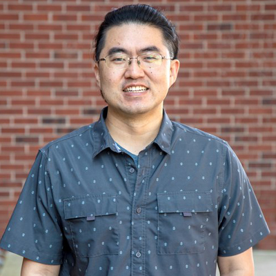 A professor smiles while posing for a headshot outside in front of a brick wall.