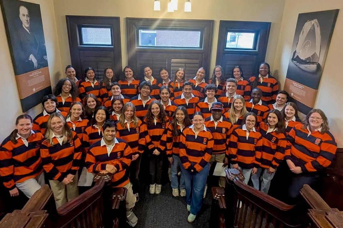 The image shows a group of people standing on a staircase, posing for a photo. They are all wearing matching orange and black striped shirts. The setting appears to be indoors, with wooden paneling and framed pictures on the walls. There are two light fixtures on the ceiling above them.