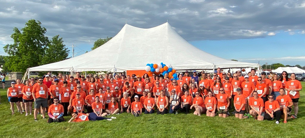 A large group of SU employees wear matching orange shirts and pose for a photo in front of a white tent on a grassy field. The sky is partly cloudy, and there are blue and orange balloons tied to the tent.