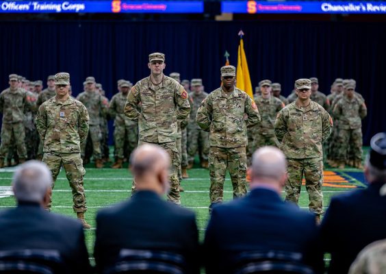 A group of military personnel stand in formation on a field. In the foreground, there are four soldiers in camouflage uniforms standing at attention with their hands behind their backs. Behind them, more soldiers are lined up in rows. The background features a dark curtain and two digital screens displaying text.