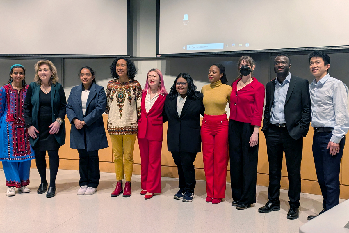 A group of ten people standing in a row in front of a classroom or lecture hall. The individuals are dressed in various styles, including traditional and formal attire. The background features a large projection screen and whiteboard.