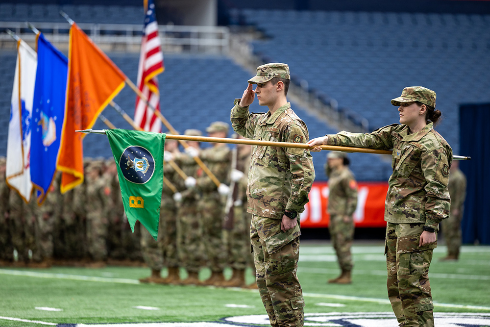 A military ceremony takes place on a field. Two soldiers in camouflage uniforms are standing in the foreground; one is saluting while the other is holding a flagpole with a green flag. In the background, more soldiers are standing in formation, and several flags, including an American flag, are visible.