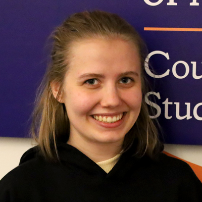 A person with shoulder-length hair is standing in front of a blue and orange sign. 