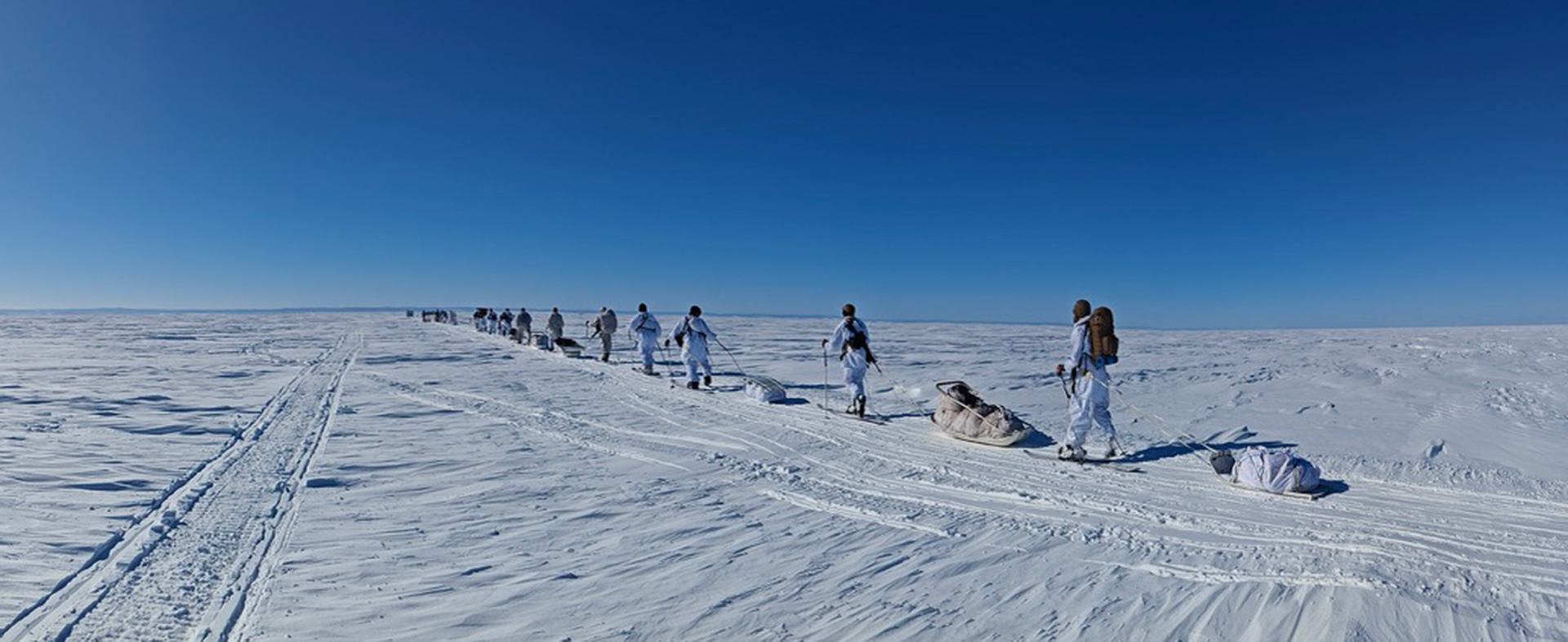 The image shows a group of people dressed in white winter gear trekking across a vast, snow-covered landscape under a clear blue sky. They are pulling sleds loaded with supplies and equipment. The scene is expansive, with the horizon stretching far into the distance, emphasizing the isolation and harshness of the environment.