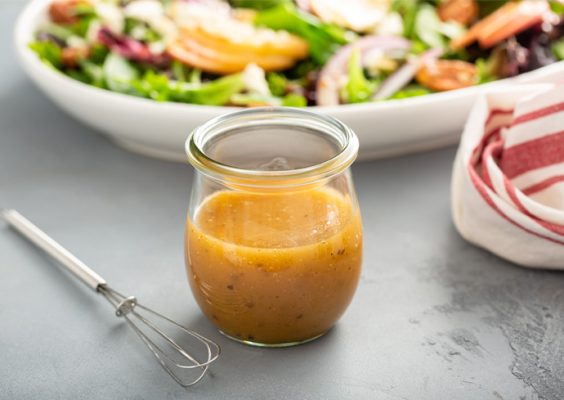 A small glass jar filled with salad dressing is placed on a table. To the left of the jar, there is a small whisk. In the background, there is a white bowl containing a colorful salad with various vegetables and greens. A red and white striped cloth napkin is partially visible on the right side of the image.
