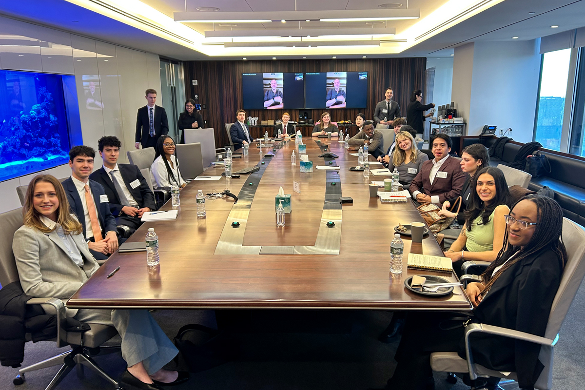 The image shows a large conference room with a long, rectangular wooden table surrounded by people in business attire. The room has modern decor with a large fish tank on the left wall and two screens at the far end displaying a video call. There are water bottles, notebooks, and name tags on the table. Several people are standing at the back of the room. The room has large windows allowing natural light to enter.