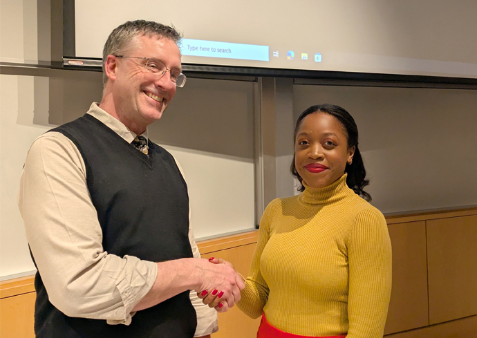 A man and a woman are shaking hands in what appears to be a classroom or lecture hall. The man is wearing glasses, a beige shirt, and a black vest. The woman is wearing a yellow sweater and red skirt. There is a projection screen in the background.