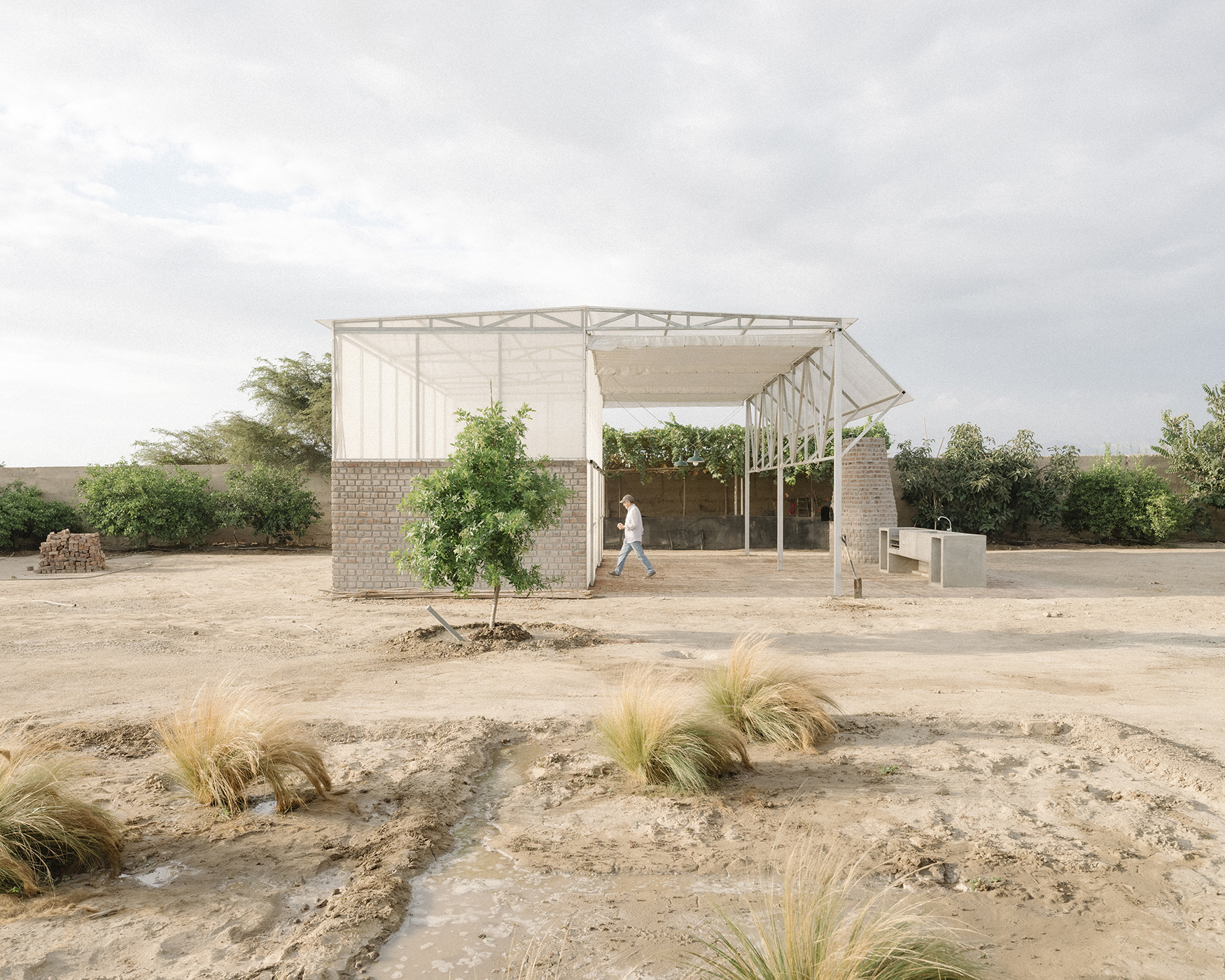 A partially constructed building with an open framework. The structure has a metal roof supported by white beams and columns. There are some brick walls on the sides, but the front is open, revealing a person walking inside. In the foreground, there is dry soil with patches of grass and small plants. The background includes trees and shrubs under a cloudy sky.