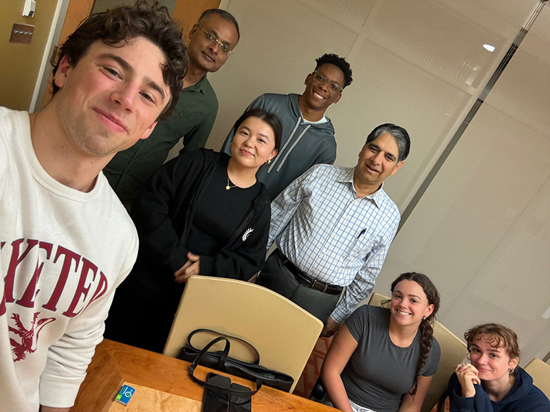 A group of eight people are gathered in a room, with some standing and others sitting around a wooden table. The person in the foreground is taking a selfie, wearing a white sweatshirt with red text. The background shows part of the room's wall and door.