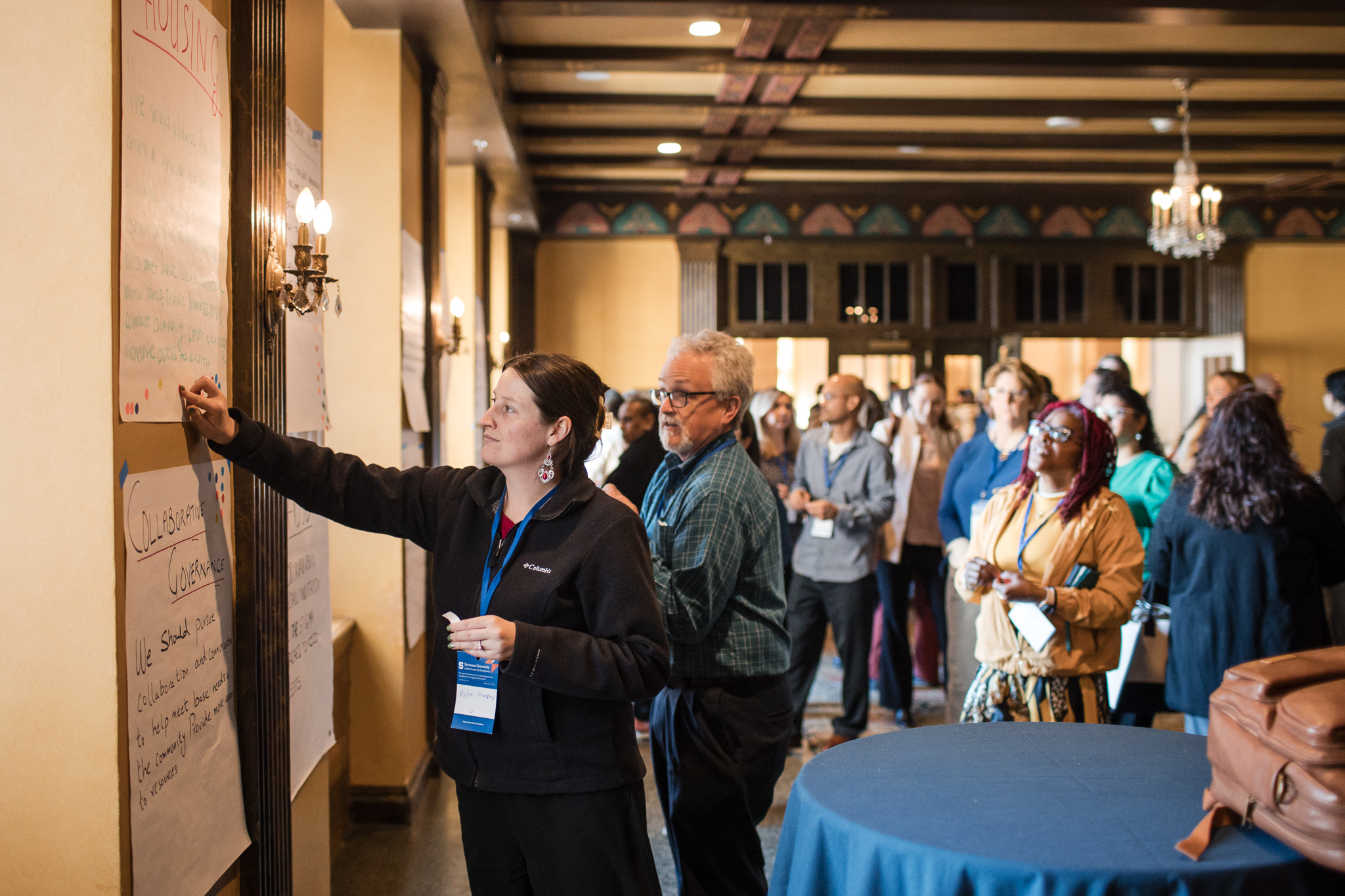 The image shows a group of people gathered in a room with ornate wooden ceilings and chandeliers. Several individuals are standing near large sheets of paper attached to the wall, which contain handwritten notes. One person is writing on one of the sheets with a marker. The room has warm lighting and a mix of casual and formal attire among the attendees. There are round tables in the foreground, one of which has a brown bag placed on it.